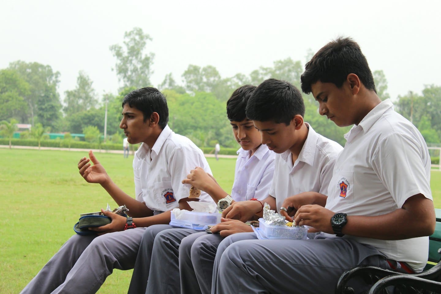 Four students in white uniforms eat lunch on a park bench.